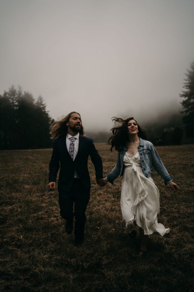 Couple running through a moody field, fog coming through the dark trees in the background. Northern California Elopement photography in the redwoods. 