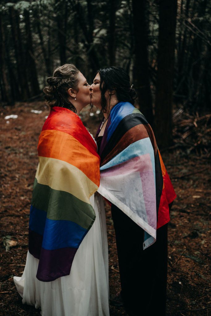Lesbian Couple kissing with the LGBTQ+ flag wrapped around them celebrating their marriage and marriage equality. They are surrounded by a rainy forest near Lake Tahoe California for their intimate wedding with close friends.