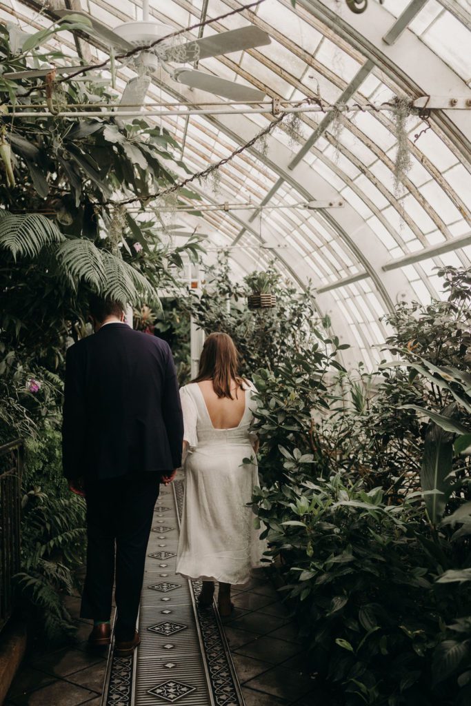 Couple walking away from the camera in a green house surrounded by incredible plants with light streaming through the windows. San Francisco Elopement Photography