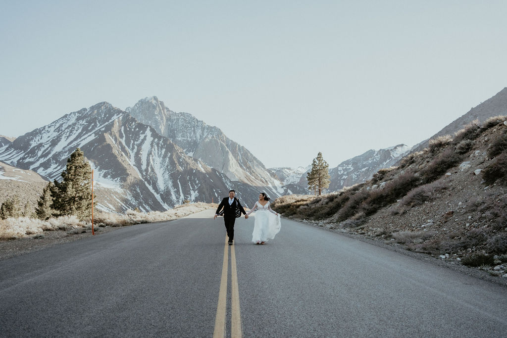 Eloping Couple running down a slopped road that looks like it drives into a mountain looming massive in the background. A few trees dot the side of the road during their adventure elopement in Mammoth Lakes California with their elopement photographer.