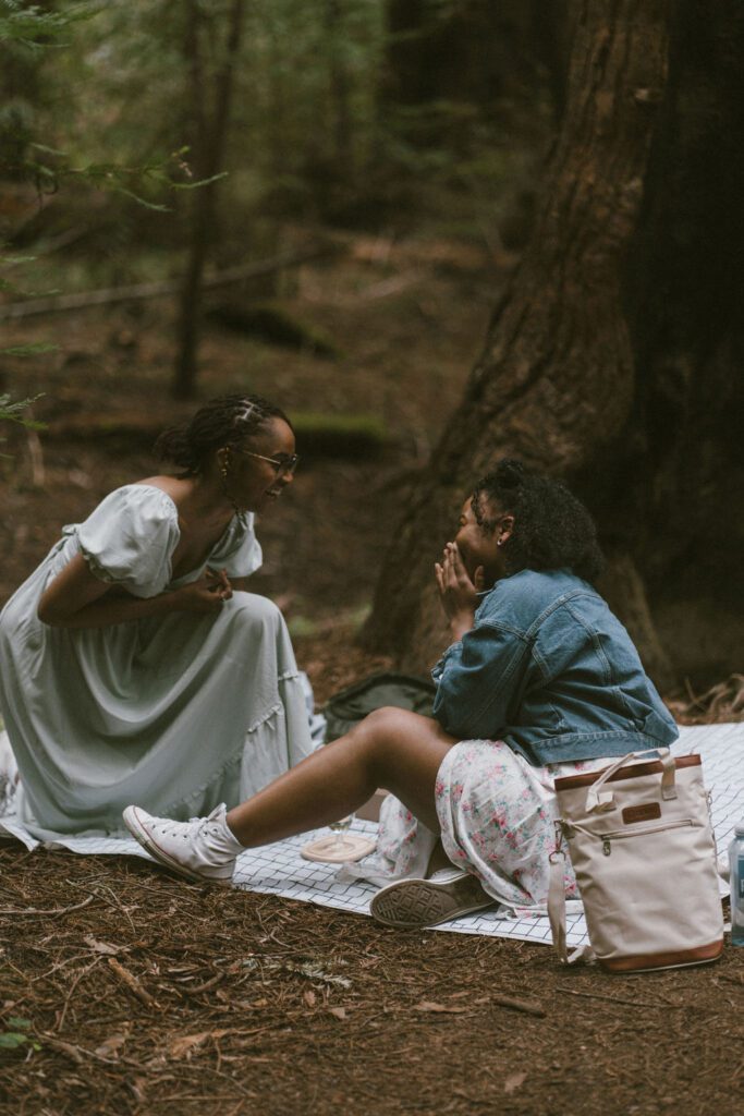 One girl in a sage blue dress on her knees proposing to another girl in a white dress and Jean jacket on a picnic blanket in the Redwoods, Engagement photos.