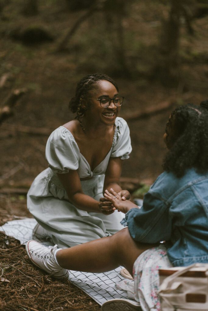 A girl proposing to her girlfriend putting a ring on her finger and smiling on a picnic blanket. Engagement photos in the redwood.